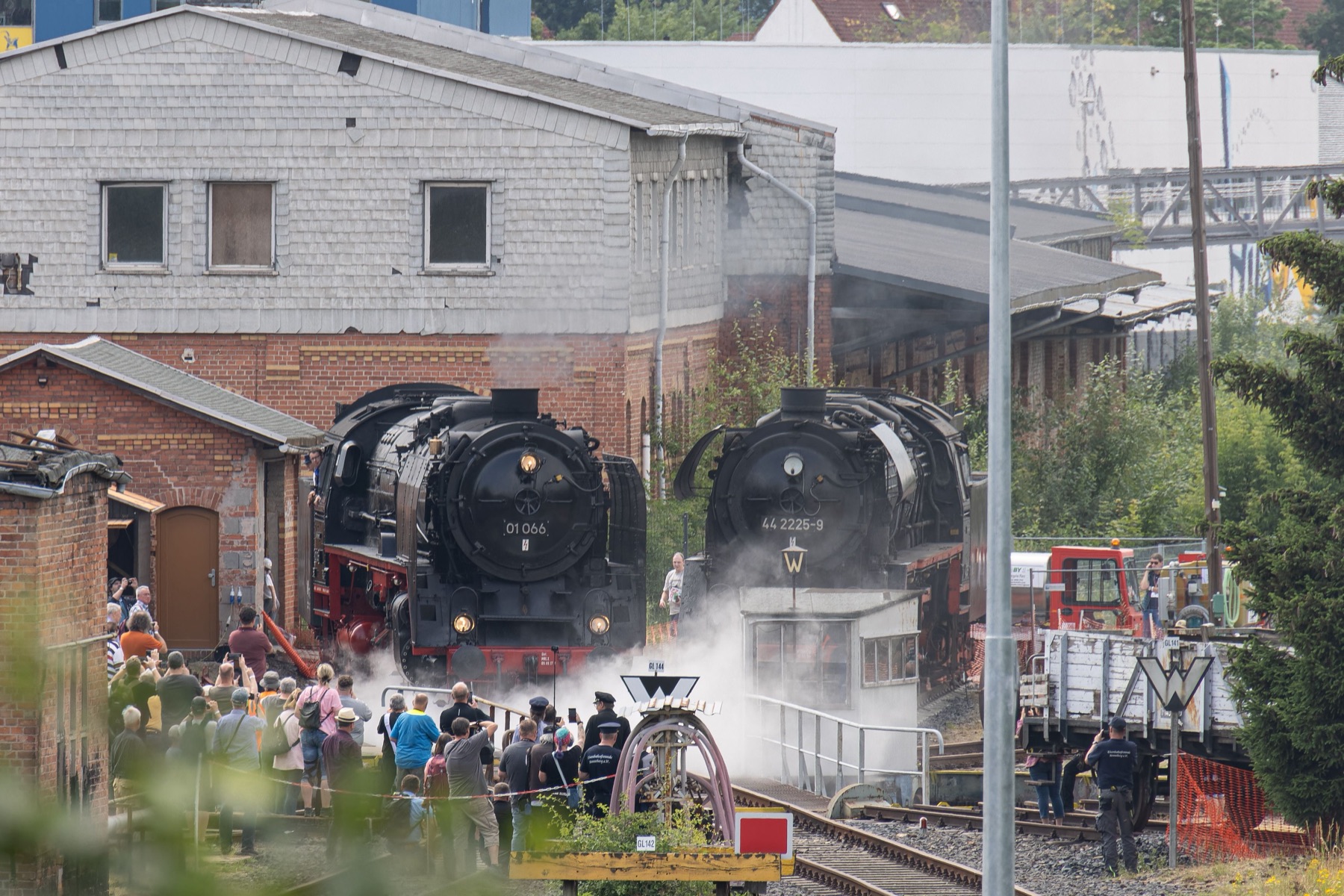 Two black steam locomotives simmering side by side outside the brick engine house while a crowd of visitors looks on